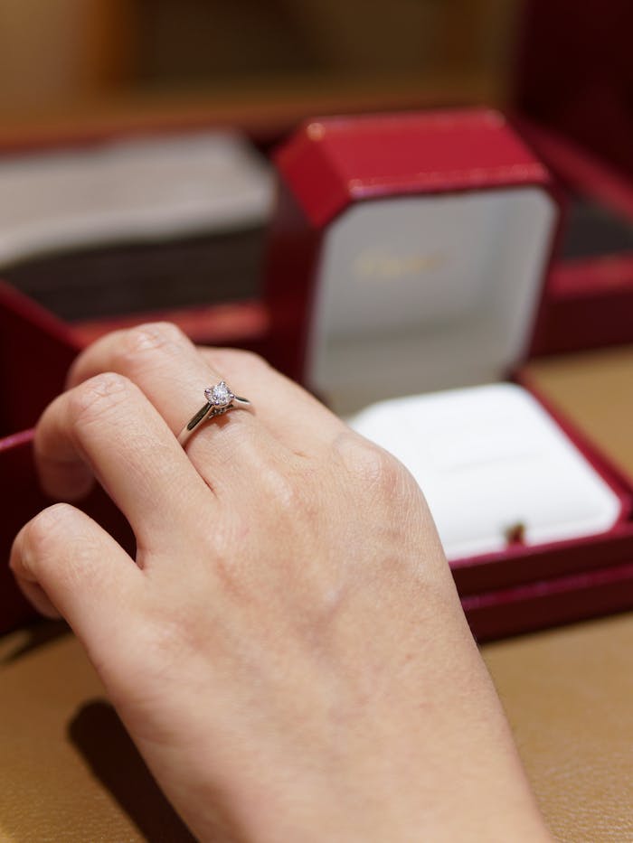 get-in-touch Close-up of a hand with an elegant diamond engagement ring against a blurred jewelry box background.